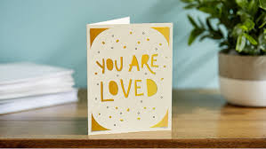 Greeting card with 'You are loved' text on a wooden surface with books and a plant in the background.